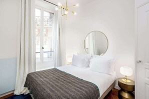Une chambre d’hôtel avec un lit, un miroir rond, une lampe de chevet et une fenêtre avec des rideaux.
