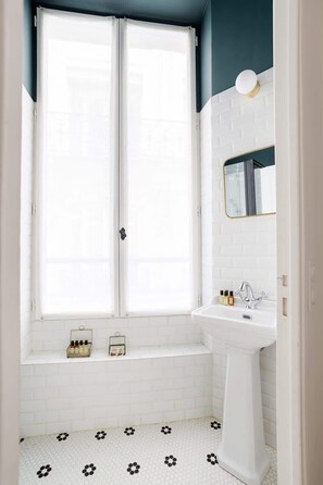 Une salle de bain avec un lavabo blanc, un miroir et une fenêtre avec des rideaux blancs.
