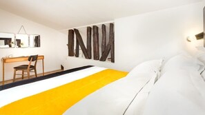 Quarto de hotel com cama, escrivaninha e cadeira. Há uma obra de arte grande na parede e um aparelho de luz sobre a cama.