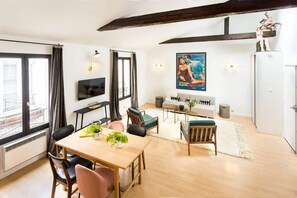 Uma sala de estar moderna com mesa de jantar de madeira, assentos confortáveis, televisão de tela plana e uma grande janela com cortinas.