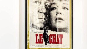 A poster of 'Le Chat' by Jean Gabin and Simone Signoret, displayed on a wall above a wooden dining table with chairs.