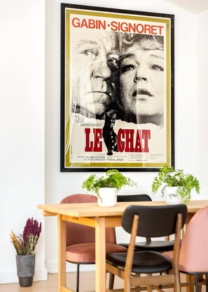 Um cartaz de 'Le Chat', com Jean Gabin e Simone Signoret, exposto numa parede acima de uma mesa de jantar de madeira com cadeiras.