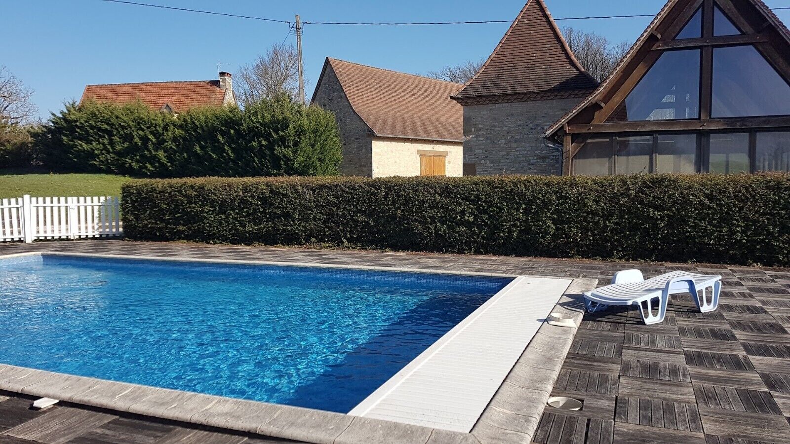 A rectangular swimming pool with a wooden deck and lounge chairs, surrounded by a hedge and a white picket fence.