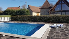A rectangular swimming pool with a wooden deck and lounge chairs, surrounded by a hedge and a white picket fence.