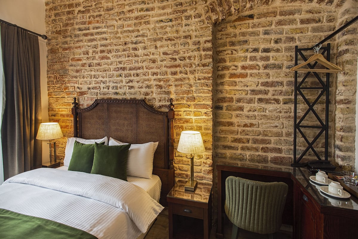 Ein Hotelzimmer mit Backsteinwand, einem hölzernen Bett mit grünen Kissen, zwei Nachttischlampen, einem Holznachttisch, einem grünen Sessel und einem kleinen Tisch mit Tassen und Tablett.