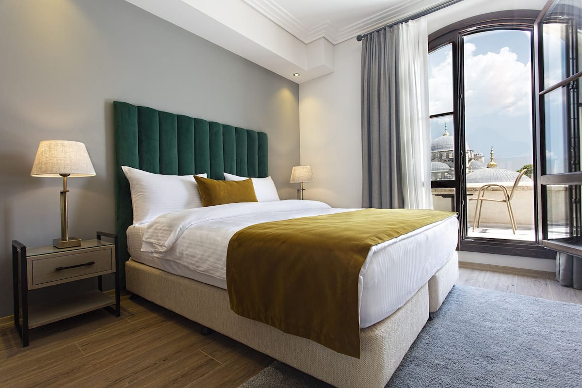 Ein Hotelzimmer mit einem großen Bett, zwei Nachttischen, Blick auf ein Gebäude durch ein großes Fenster und einem Balkon mit einem Stuhl.