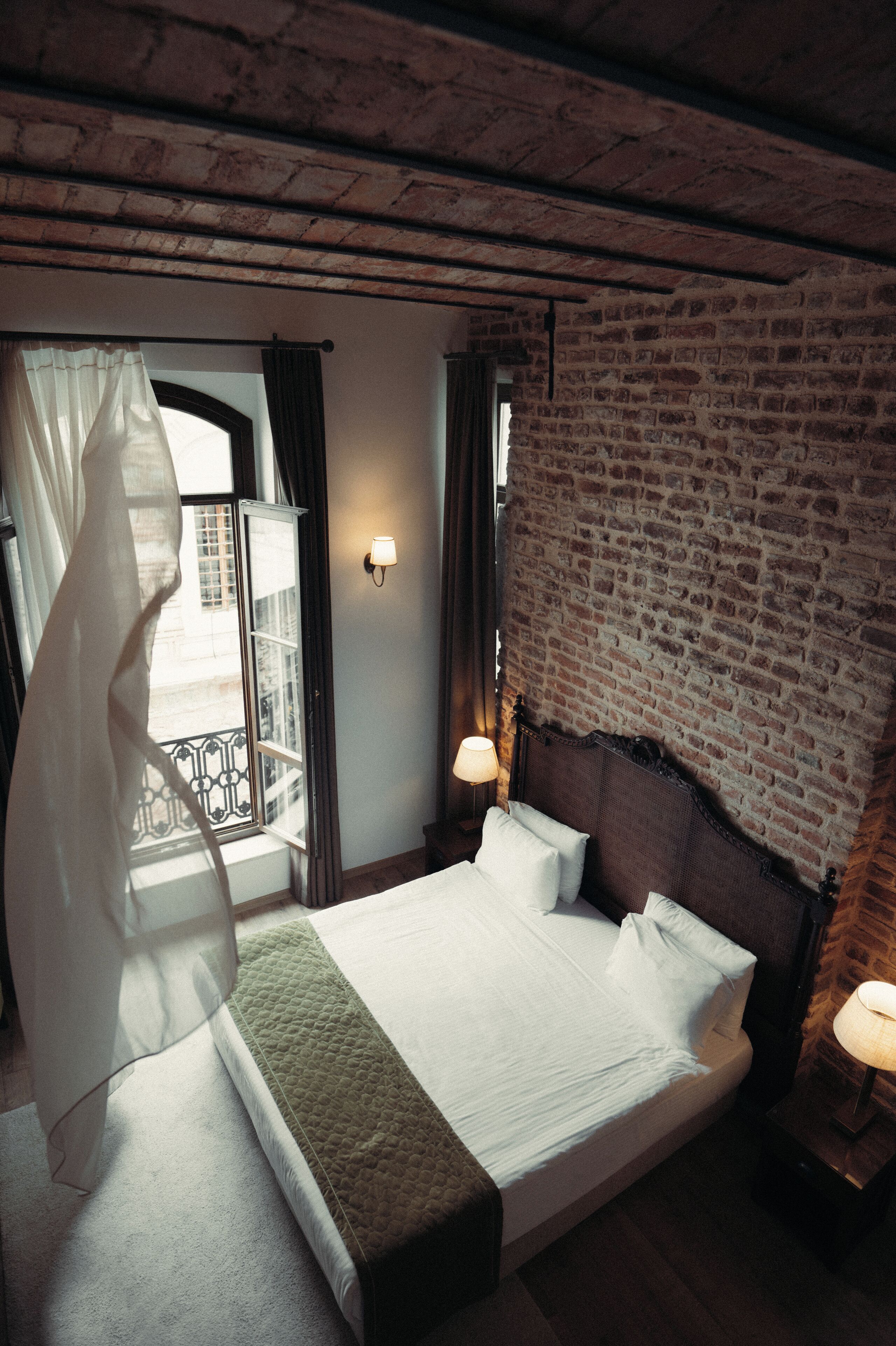Um quarto com parede de tijolos, uma cama grande com roupa de cama branca, uma mesa de cabeceira com abajur e vista pela janela com cortinas.