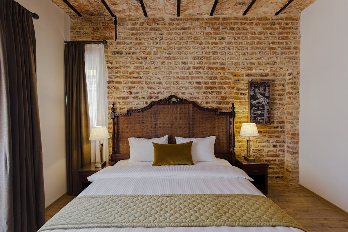 Ein Schlafzimmer mit Backsteinwand, Holz-Kopfstück, einem Bett mit weißer Bettwäsche, zwei Nachttischlampen und einem Fenster mit Vorhängen.
