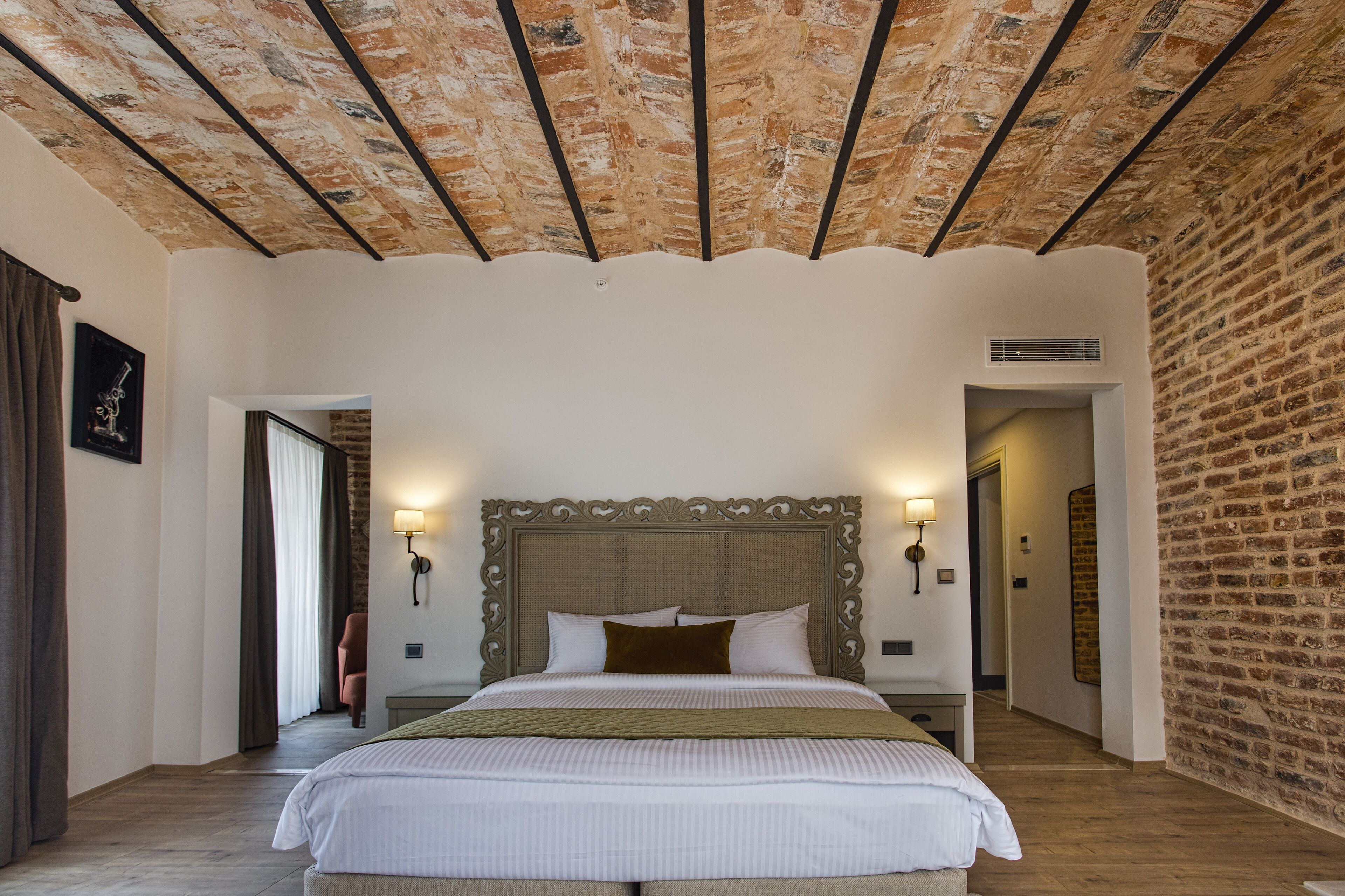 Une chambre avec un grand lit, un mur en briques, un plafond en bois et une porte donnant sur une autre pièce.