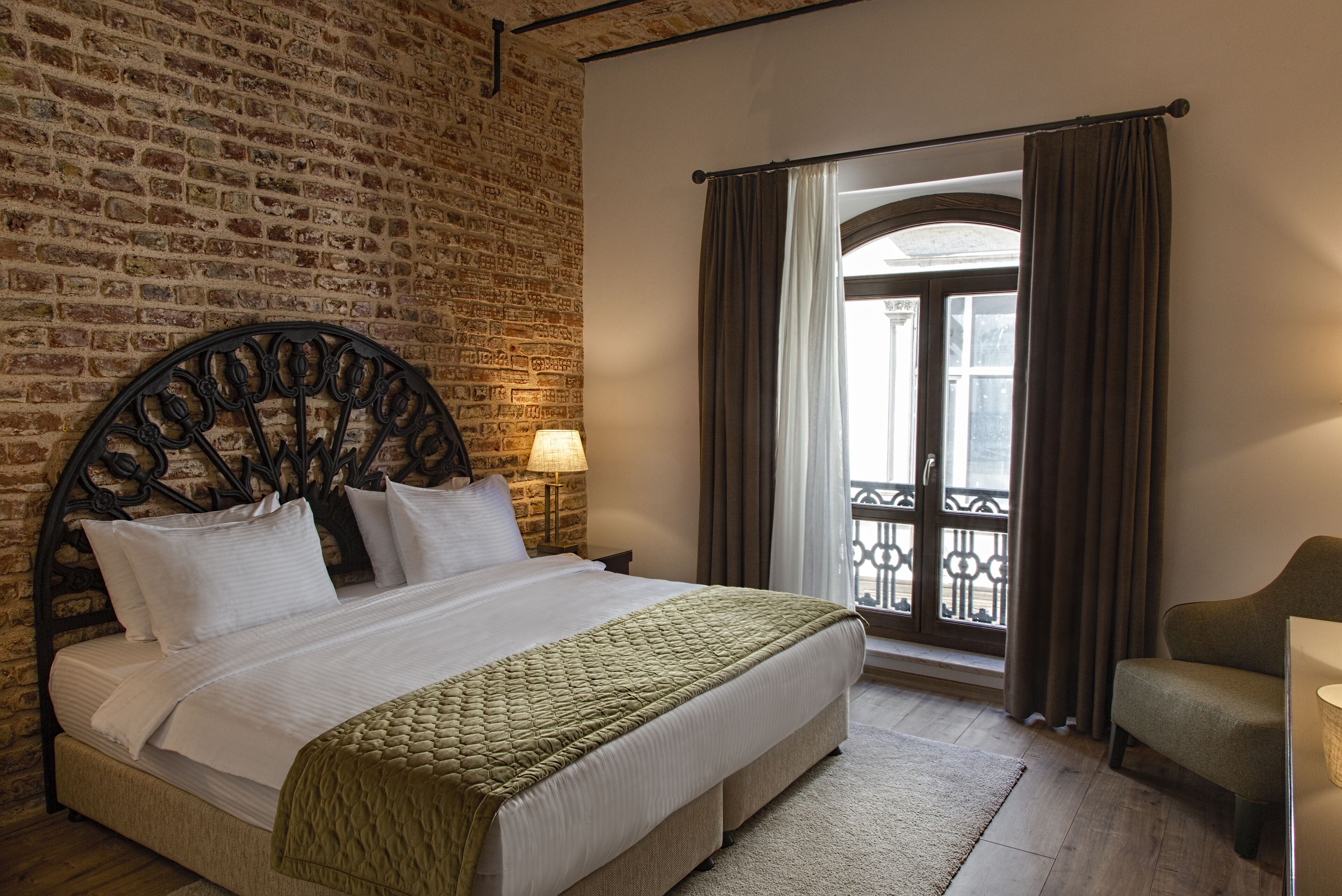 A hotel room with a brick wall, a bed with white linens, a green quilt, a nightstand with a lamp, and a view of a balcony with curtains.