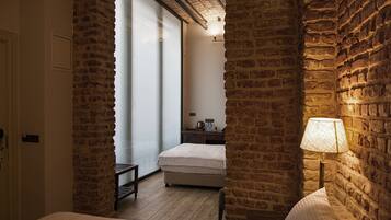 A hotel room with a brick wall, a bed, a nightstand, a lamp, and a view through a glass door.