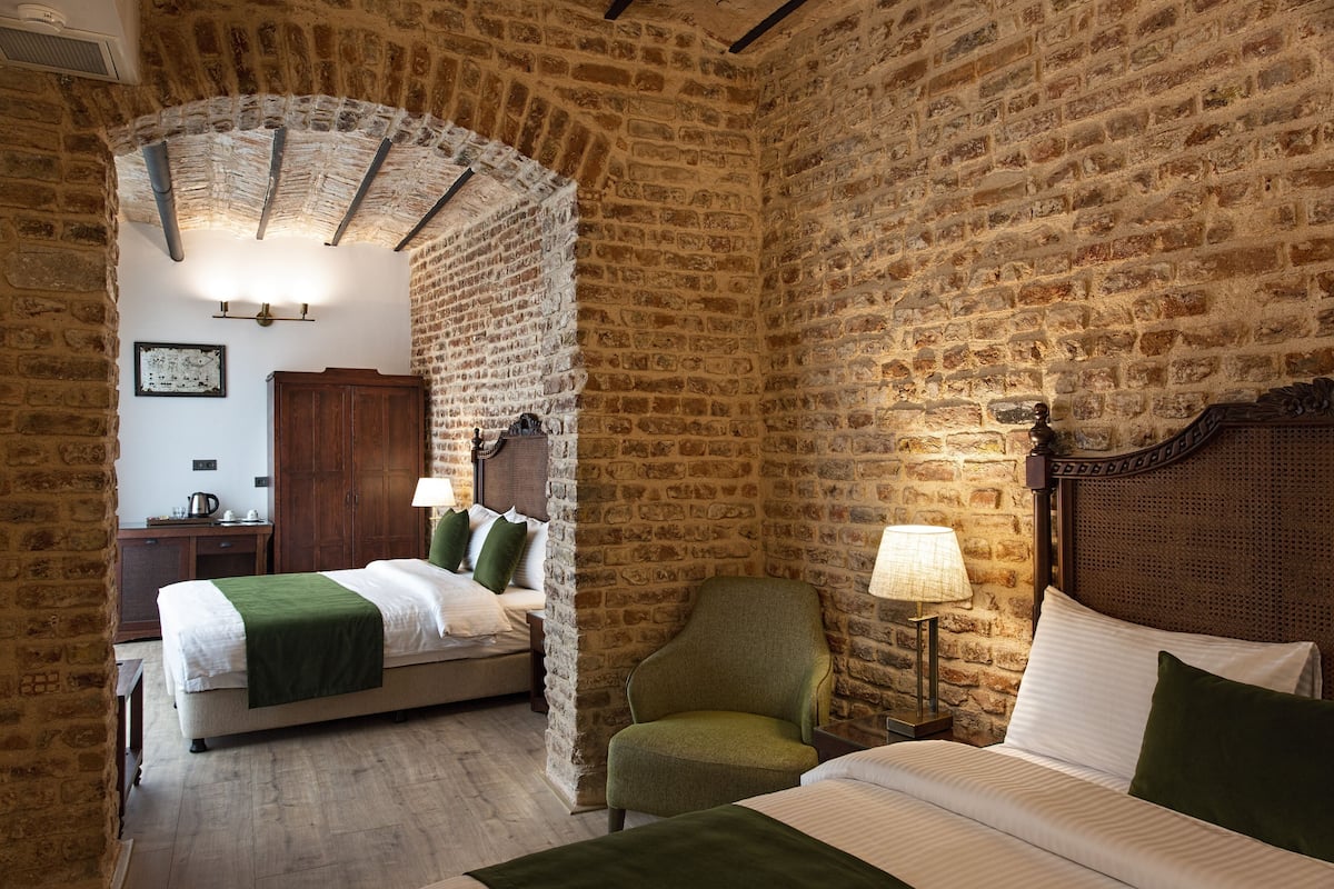 Ein Zimmer mit Steinwand, einem Bett mit grüner Decke, einem Sessel, einem Nachttisch mit Lampe und einem Holzschrank.