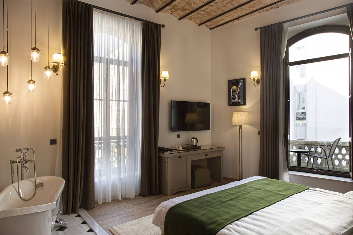 Ein Hotelzimmer mit einem großen Bett, einem Schreibtisch mit Flachbildfernseher, einer Badewanne und einem Balkon mit Blick.