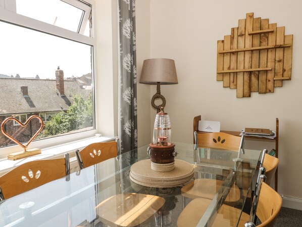 A dining area with a glass table, wooden chairs, a lamp, and a wall-mounted wooden shelf.
