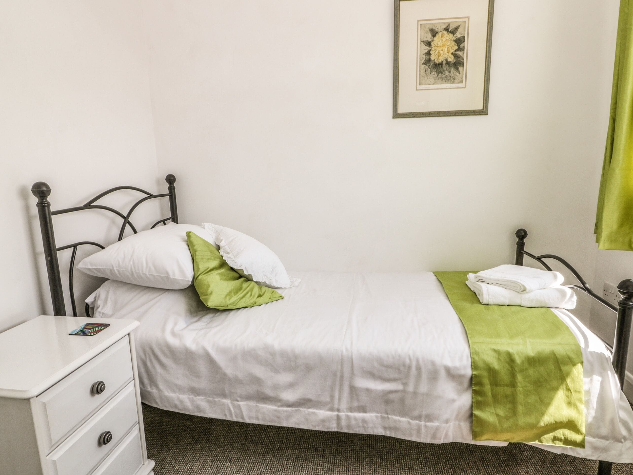 Ein ordentlich bezogenes Bett mit weißer Bettwäsche und einer grünen Decke, ein weißer Nachttisch mit Schubladen und ein gerahmtes Bild an der Wand.
