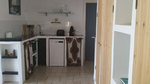 Une cuisine de taille réduite avec des meubles blancs, un plan de travail blanc et un crédence carrelée. On aperçoit une petite étagère au-dessus de l’évier, où sont posés des verres et un cadre photo. Un tapis à motifs recouvre le sol, et une porte en bois est visible sur la droite.