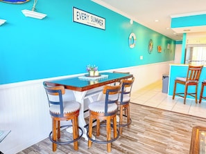 A dining area with a wooden table and chairs, a blue wall with a sign, and a kitchen in the background. - Ocean Shores 301 (North Myrtle Beach)