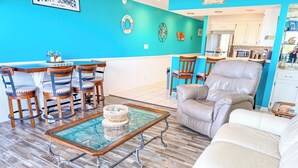 Una sala de estar con pared azul, sillas de madera, una mesa de centro de vidrio y un sofá color beige.