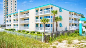 Un hotel frente a la playa con varios balcones, una decoración en blanco y azul, y una zona de playa arenosa al frente.
