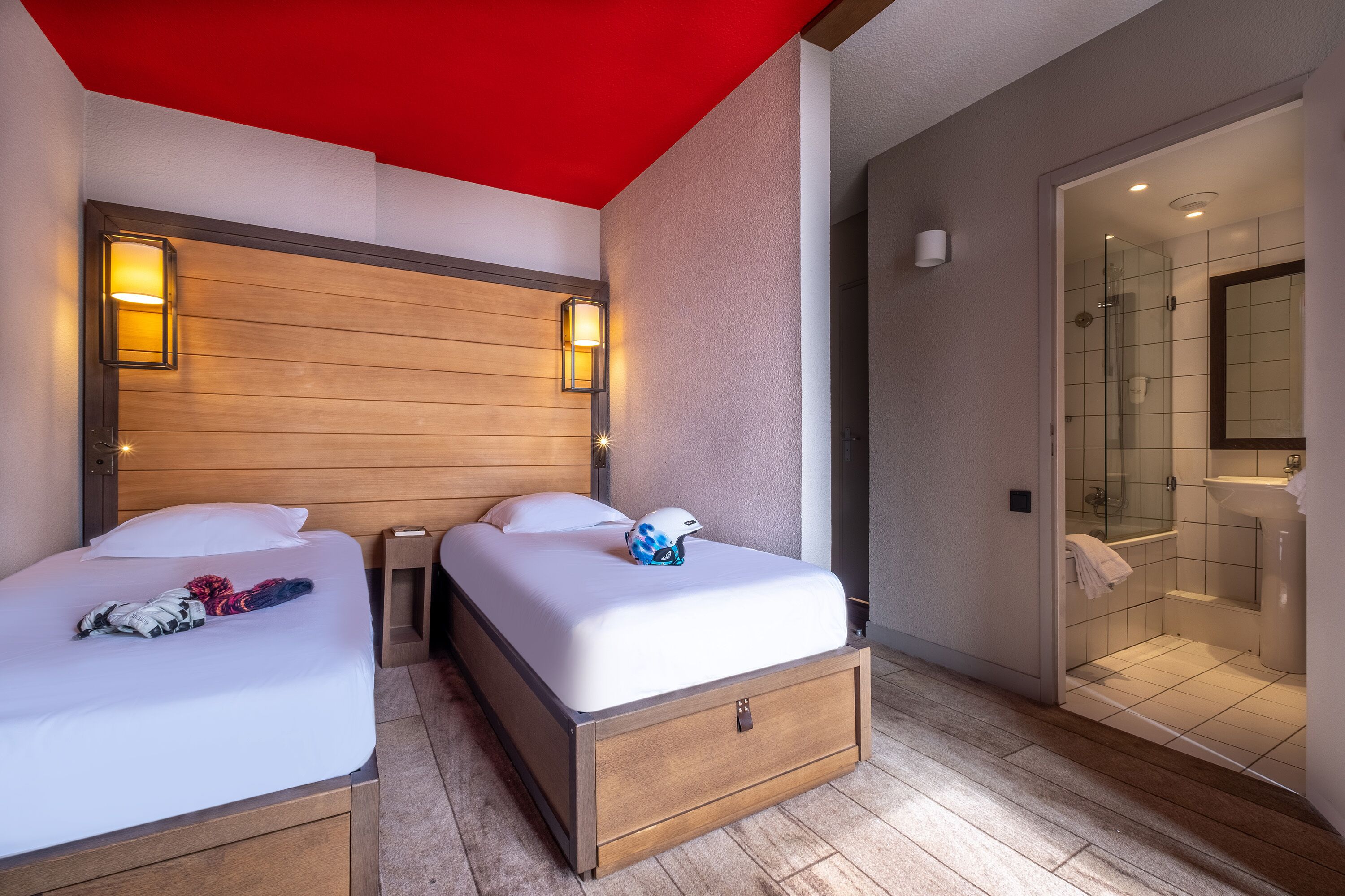 A hotel room with two beds, a wooden headboard, a red ceiling, and a glass shower enclosure.