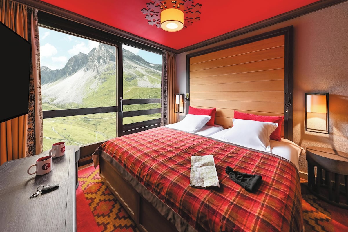 A hotel room with a large window offering a mountain view, a bed with a plaid bedspread, a bedside table with a lamp, and a TV on the left wall.