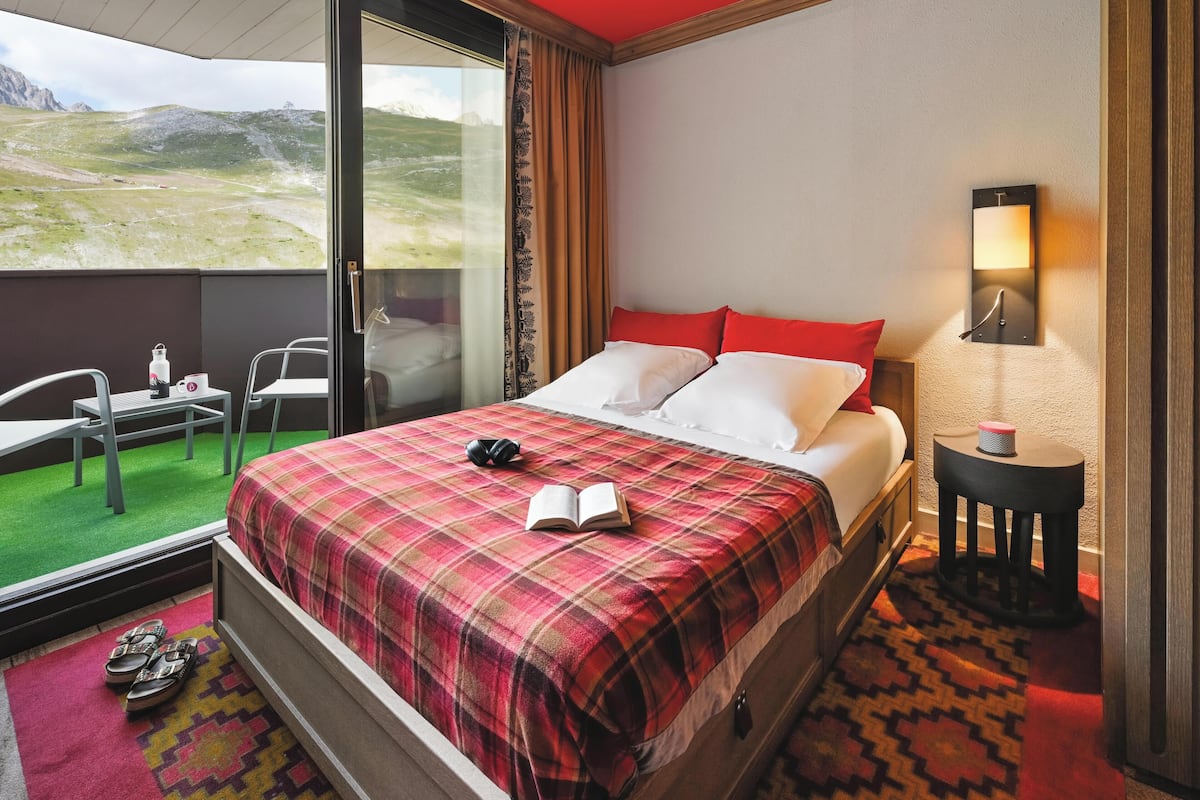 A hotel room with a bed, a small table with a chair, a lamp, and a view of a mountainous landscape.
