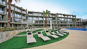Et hotel med flere balkoner, en swimmingpool og solsenge arrangeret i rækker.