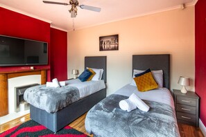 A hotel room with two beds, a flat-screen TV, a ceiling fan, and a cityscape painting on the wall. - 4br-7guests-freeparking-garden-wifi (Derby)