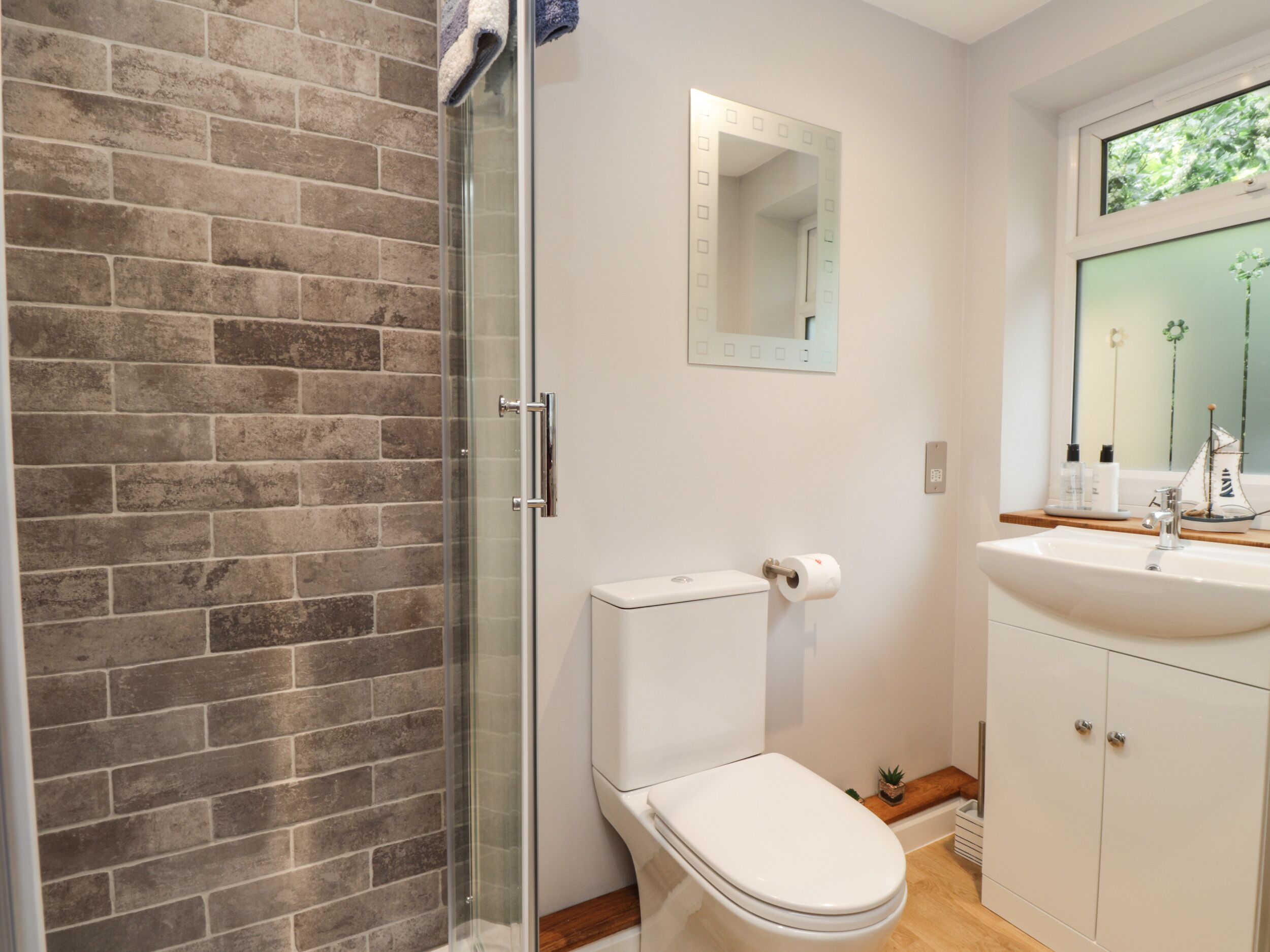 A bathroom with a glass shower enclosure, a white toilet, a sink with a mirror above it, and a window with a view of greenery.