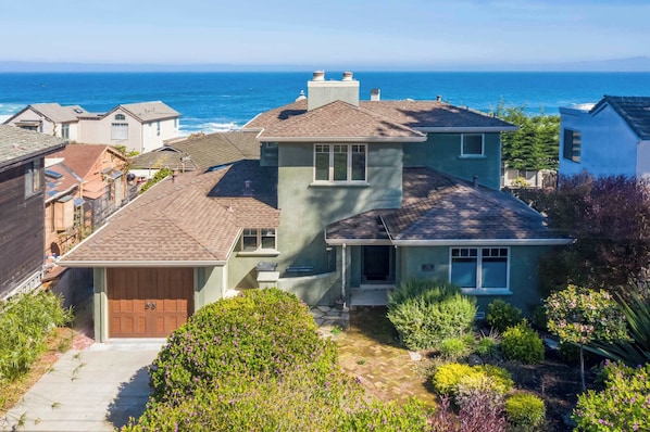 A two-story house with a garage, a front yard with various plants, and a view of the ocean.