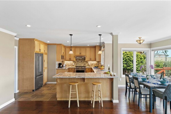 A modern kitchen with wooden cabinets, a granite countertop, and a dining area with a table set for four. - Shell's Treasure: Fully Updated Three bedroom home with Ocean Views (Pacific Grove)