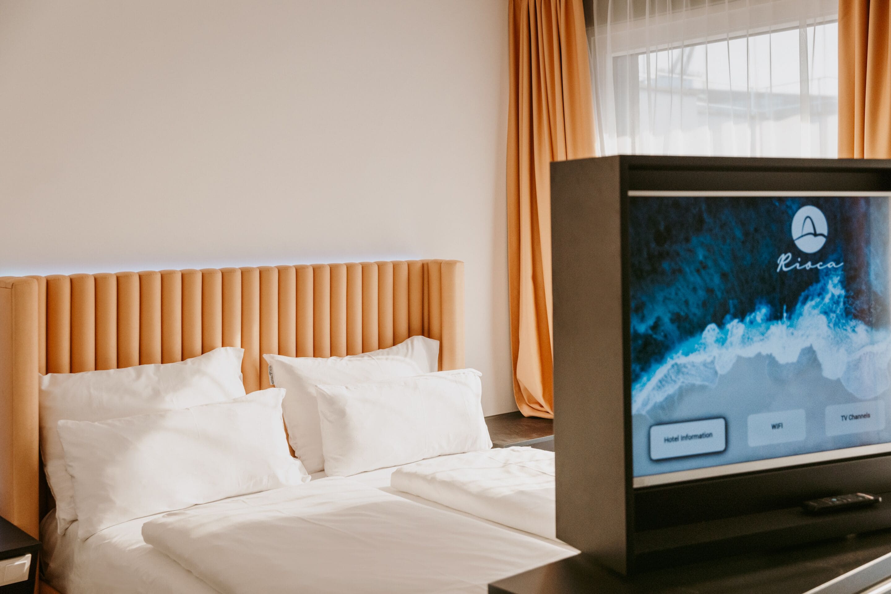Una camera d'albergo con un letto, una televisione che mostra un canale informativo dell'hotel e tende arancioni.