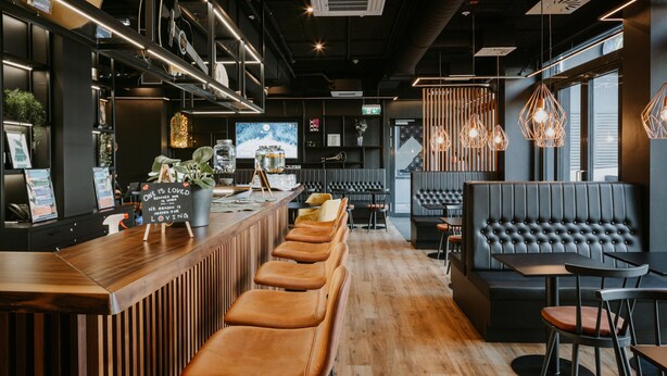 A modern bar area with wooden bar counters, leather bar stools, and a seating area with a black sofa and tables. - Rioca Vienna Posto 2 (Vienna)