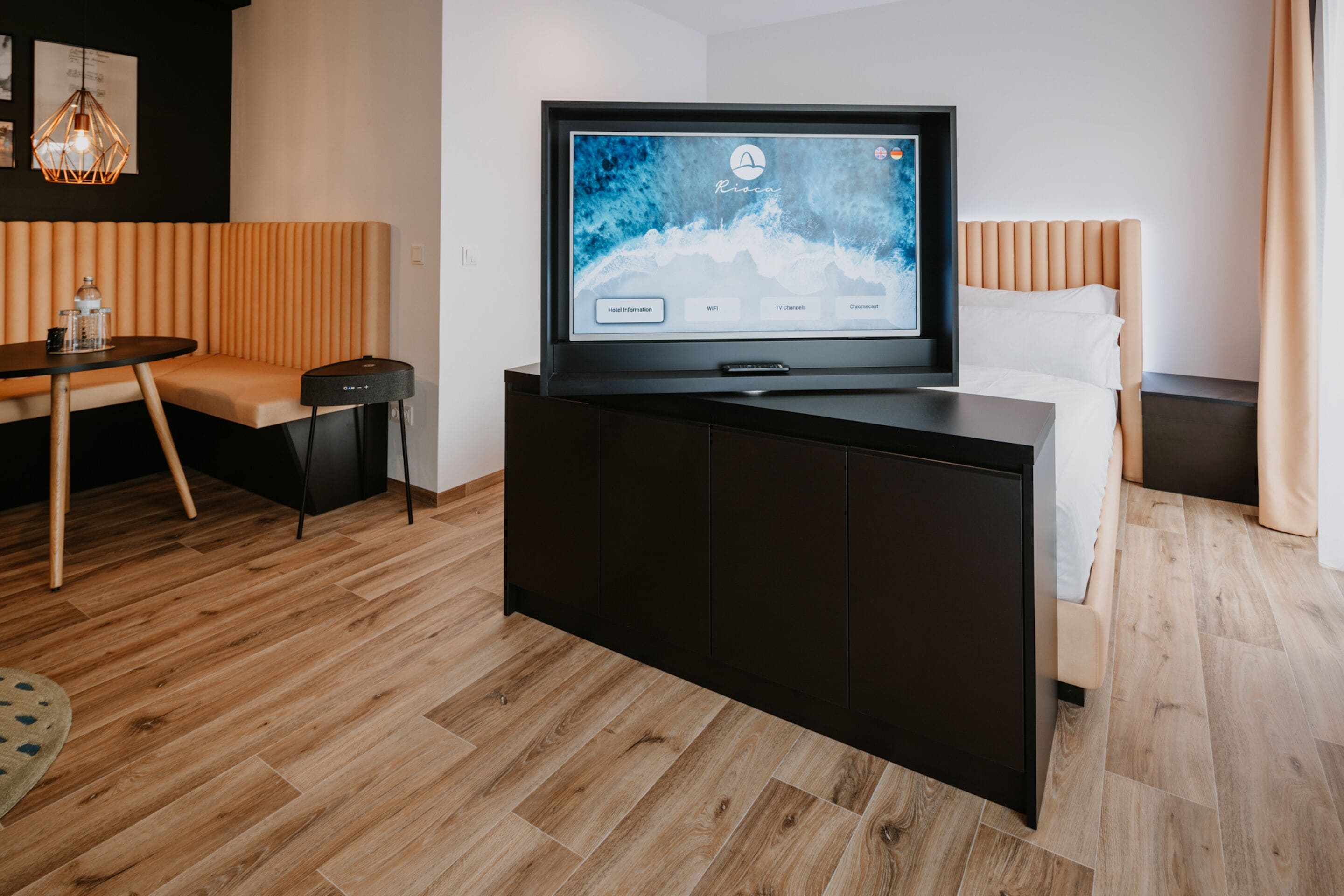 A modern hotel room with a flat-screen TV on a black stand, a wooden floor, a sofa, and a bed with white bedding.