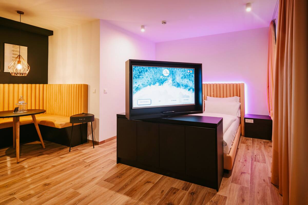 Quarto de hotel moderno com televisão de tela plana, sofá, mesa pequena e cama com cabeceira.