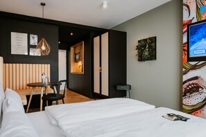 A modern hotel room with a bed, a small table with a chair, a wooden bench, and a TV on the wall.