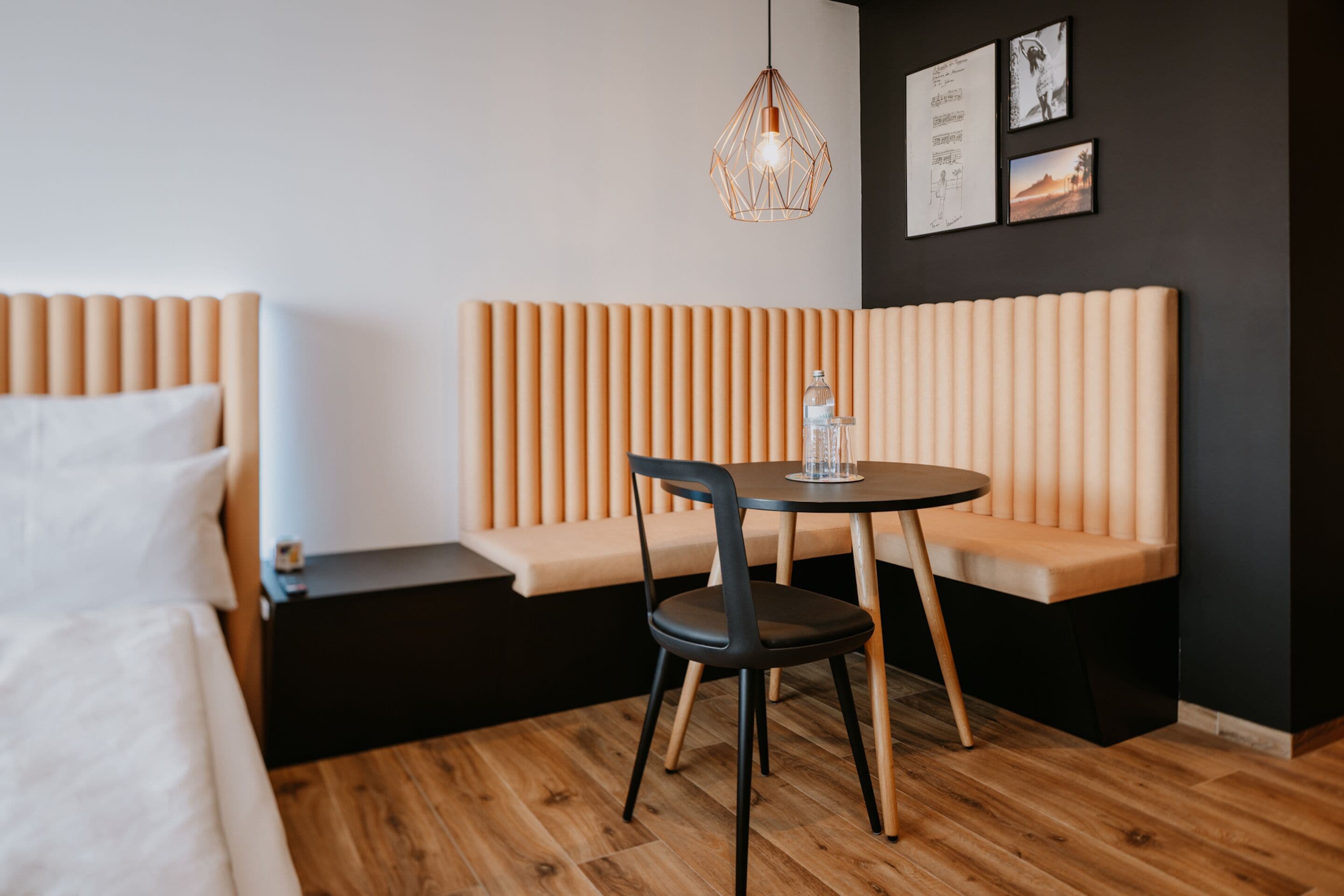 A modern hotel room with a wooden floor, a bed with white bedding, a wooden bench with a black chair, a round table with a glass bottle, and a pendant light.