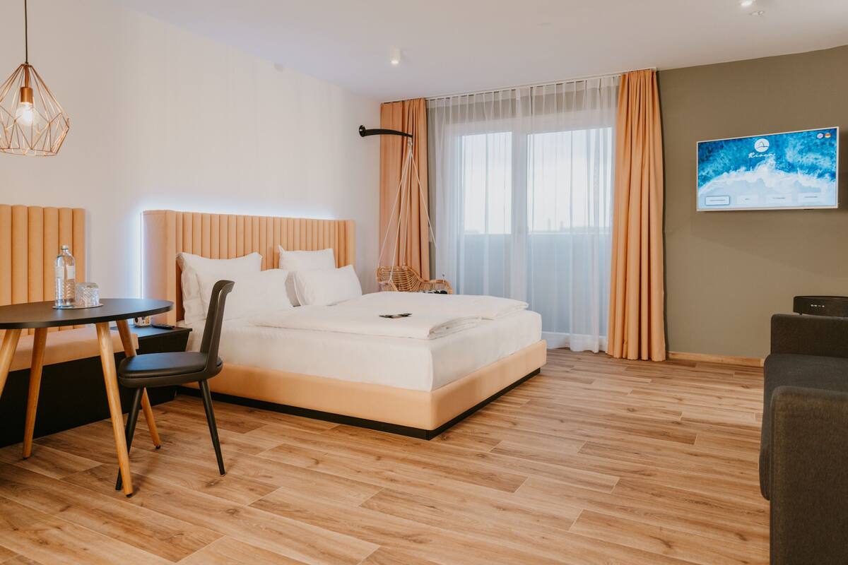 Quarto de hotel moderno com uma cama grande, área de refeições com mesa e cadeiras, televisão na parede e uma janela ampla com cortinas.