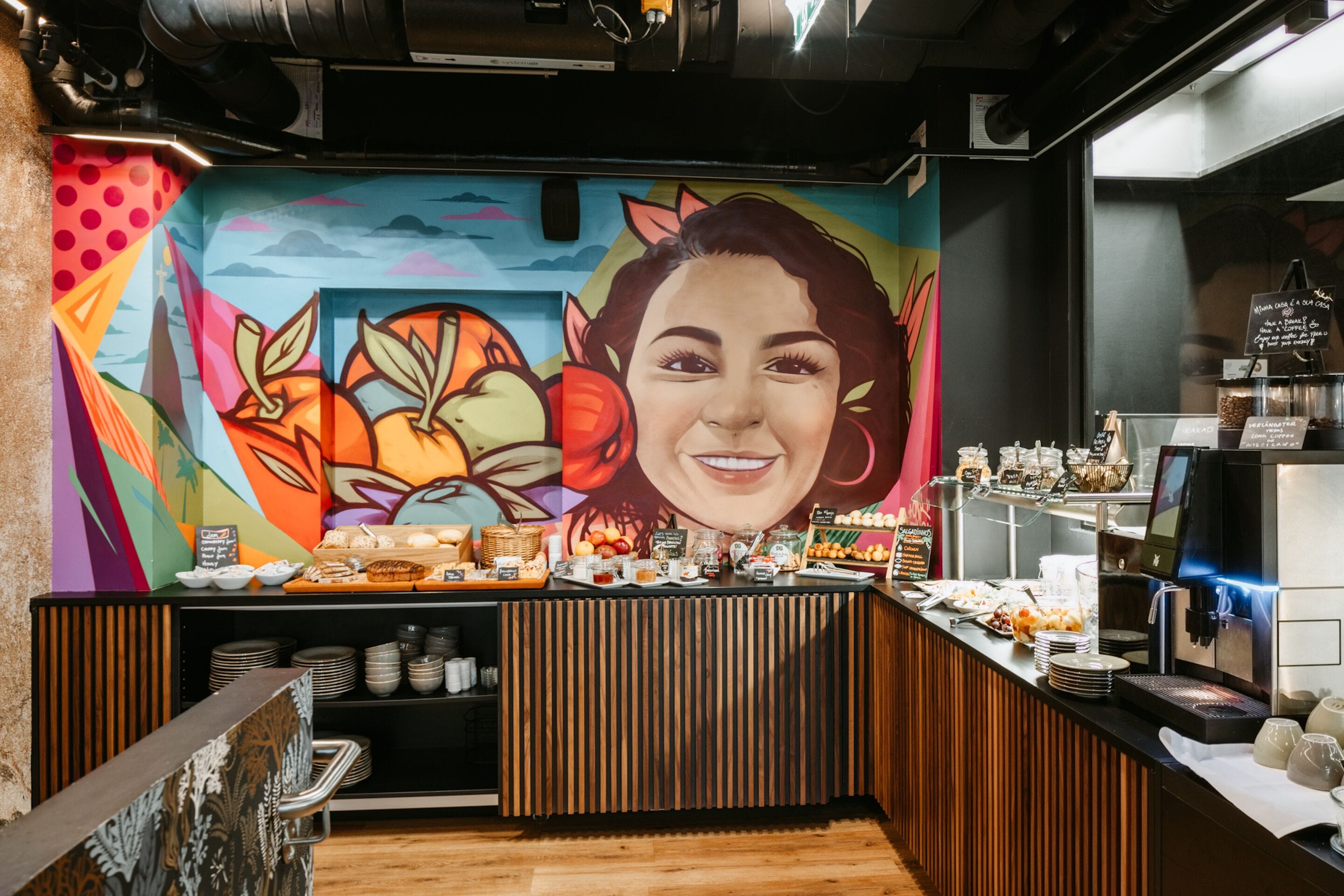 Um balcão de café da manhã com um mural colorido, uma máquina de café e diversas opções de comida.