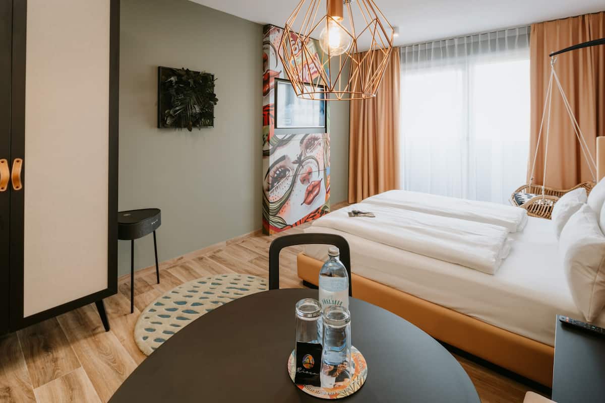 Quarto de hotel com cama, uma mesa com garrafa e copos, uma cadeira, luminária pendente e um quadro emoldurado e um adorno na parede.