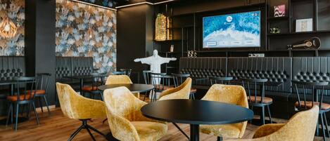 Zona de bar moderna con mesas redondas, sillas tapizadas en amarillo, una pantalla grande mostrando un paisaje montañoso y una escultura decorativa blanca.
