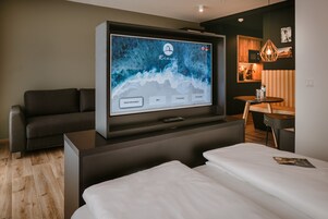 Een moderne hotelkamer met een grote flatscreen-tv waarop een hotelinterface te zien is, een bank, een bed en een klein tafeltje met een lamp.