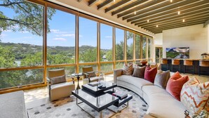 A modern living room with large windows, a sofa, a coffee table, and a view of a natural landscape. - Above Waterfalls I Premier | Party Dock I Pool I Theater I Luxury I Services (Austin)
