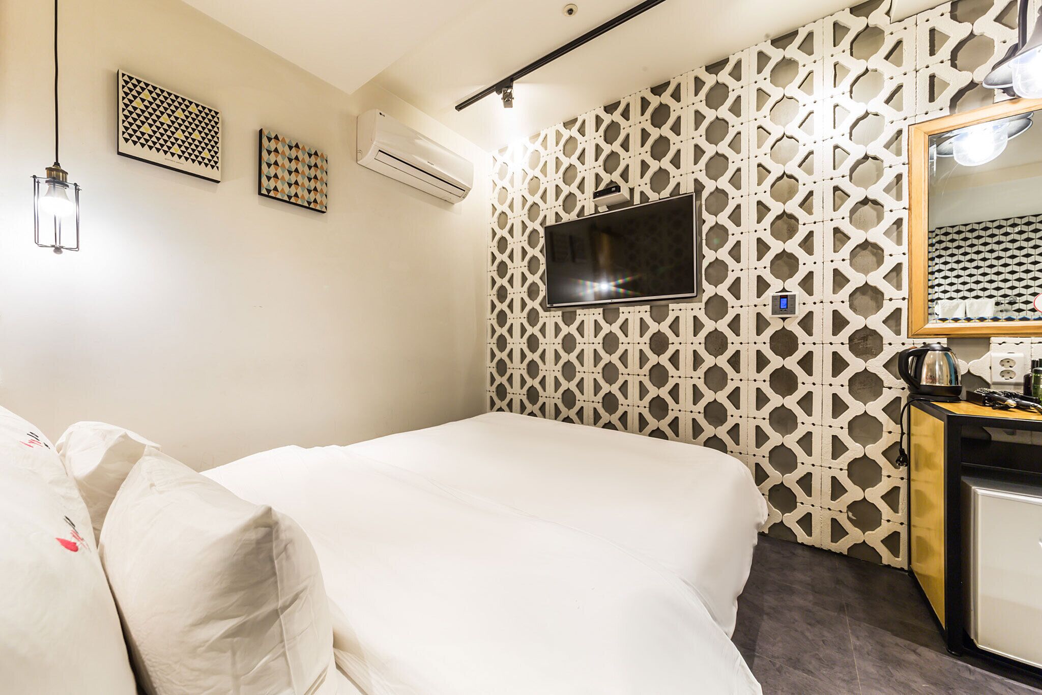Habitación de hotel con cama, televisor montado en una pared con diseño, unidad de aire acondicionado y una pequeña zona de kitchenette.