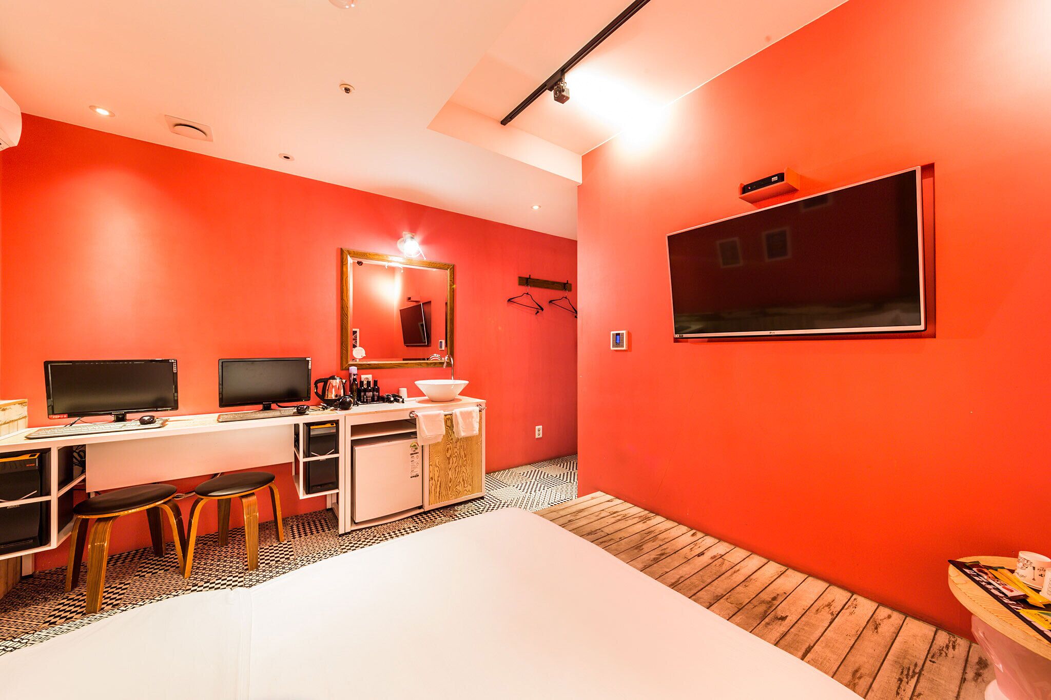 Una habitación con una pared roja, un escritorio blanco con dos monitores, un espejo y una pequeña kitchenette.