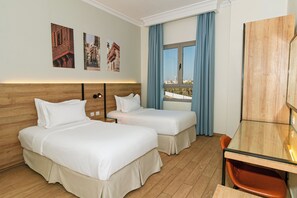 A hotel room with two beds, a desk, a chair, and a window with a view.