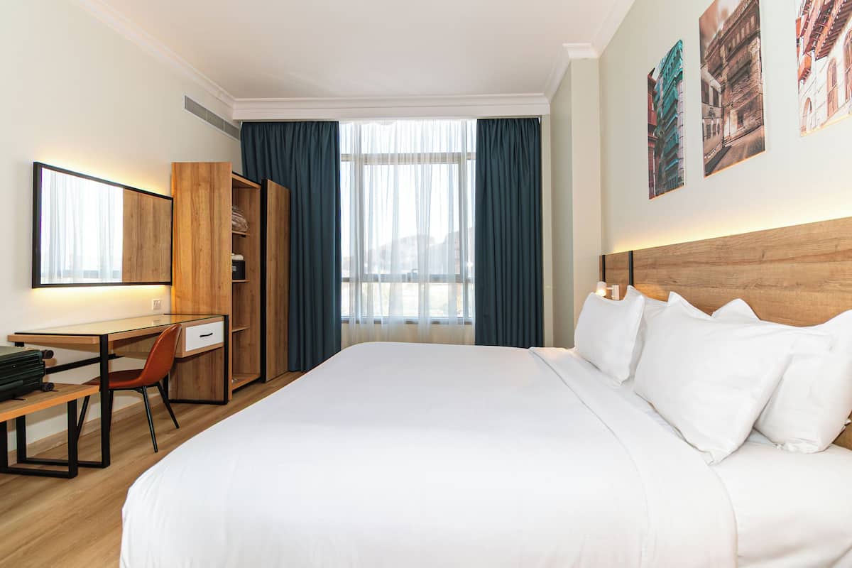 A hotel room with a bed, a desk, a chair, a wardrobe, and a large window with curtains.