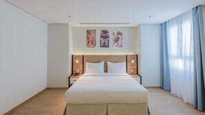 A modern hotel room with a large bed, wooden headboard, two bedside tables with lamps, and a view of the city through sheer curtains. - Hasanah Suites for Serviced Apartments. (Madinah)