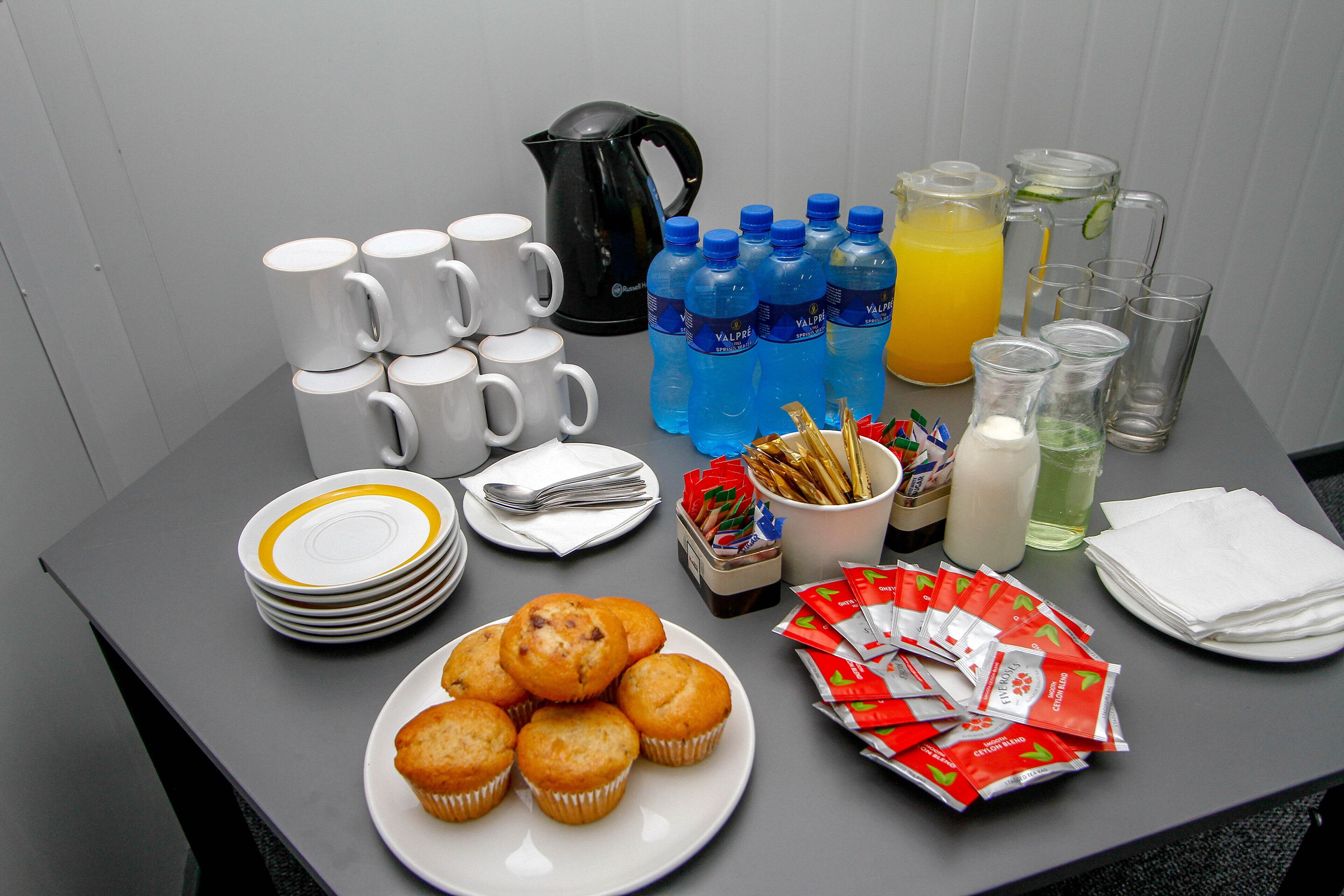 Une table de petit-déjeuner avec des tasses à café, une bouilloire, du jus et des muffins.