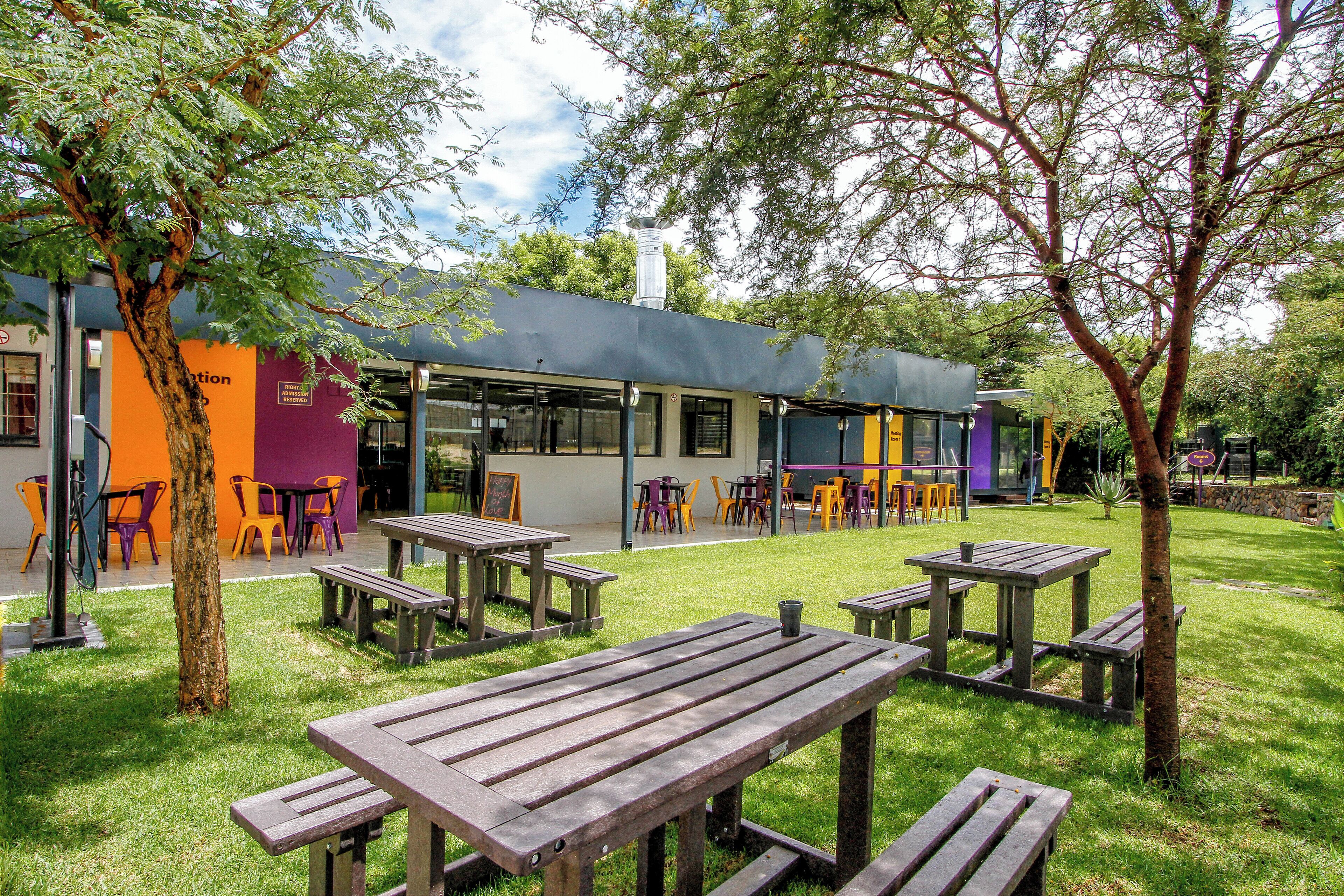 Un espace extérieur aménagé avec des tables et des chaises de pique-nique, un bâtiment doté de grandes fenêtres et un arbre au premier plan.